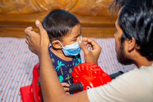 A young man is adjusting a baby's protective surgical face mask on the child's face for COVID 19 safety issue.